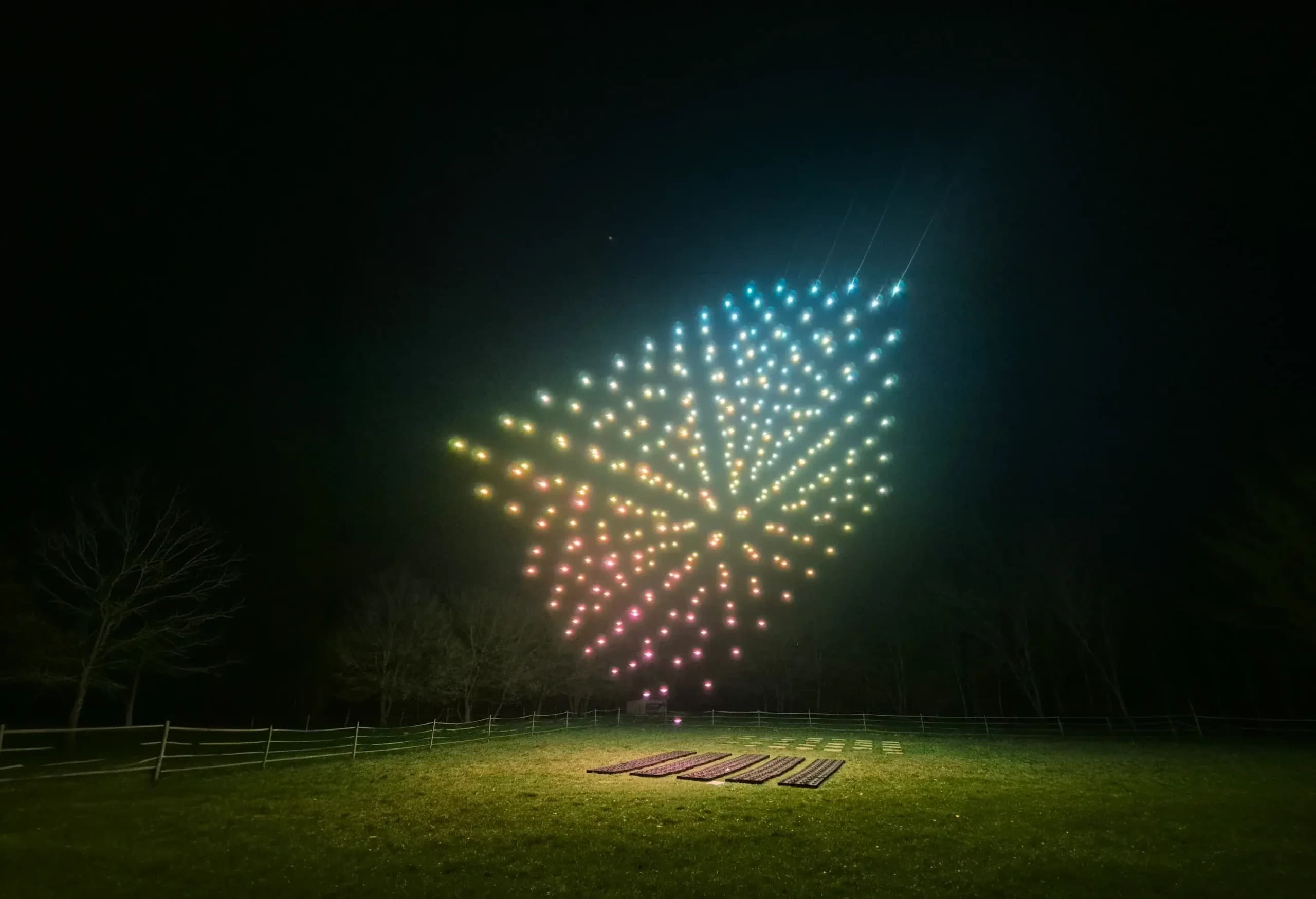 A synchronized swarm of illuminated drones forms a vibrant, multicolored geometric pattern in the night sky above a grassy field, with trees and ground equipment visible below.