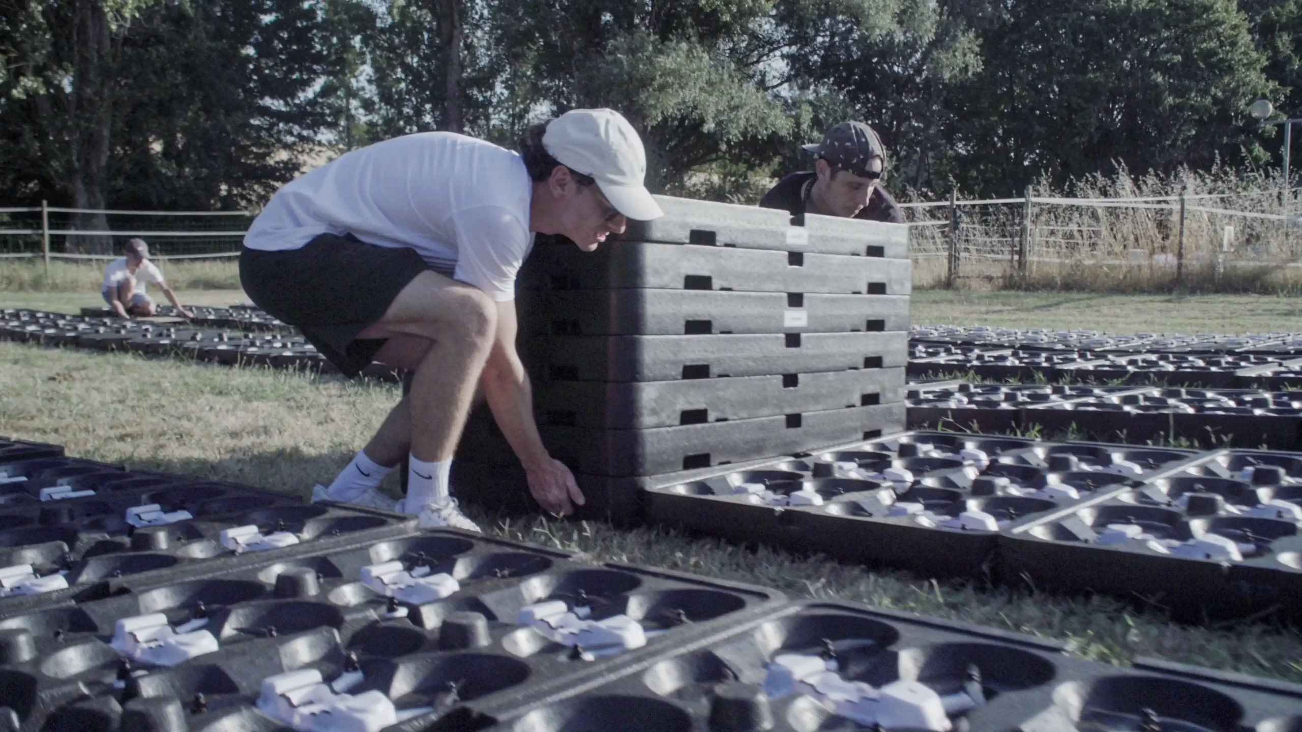 Two Drotek team members lifting stacked drone landing pad trays in a grass field, preparing the launch grid for a drone light show setup.